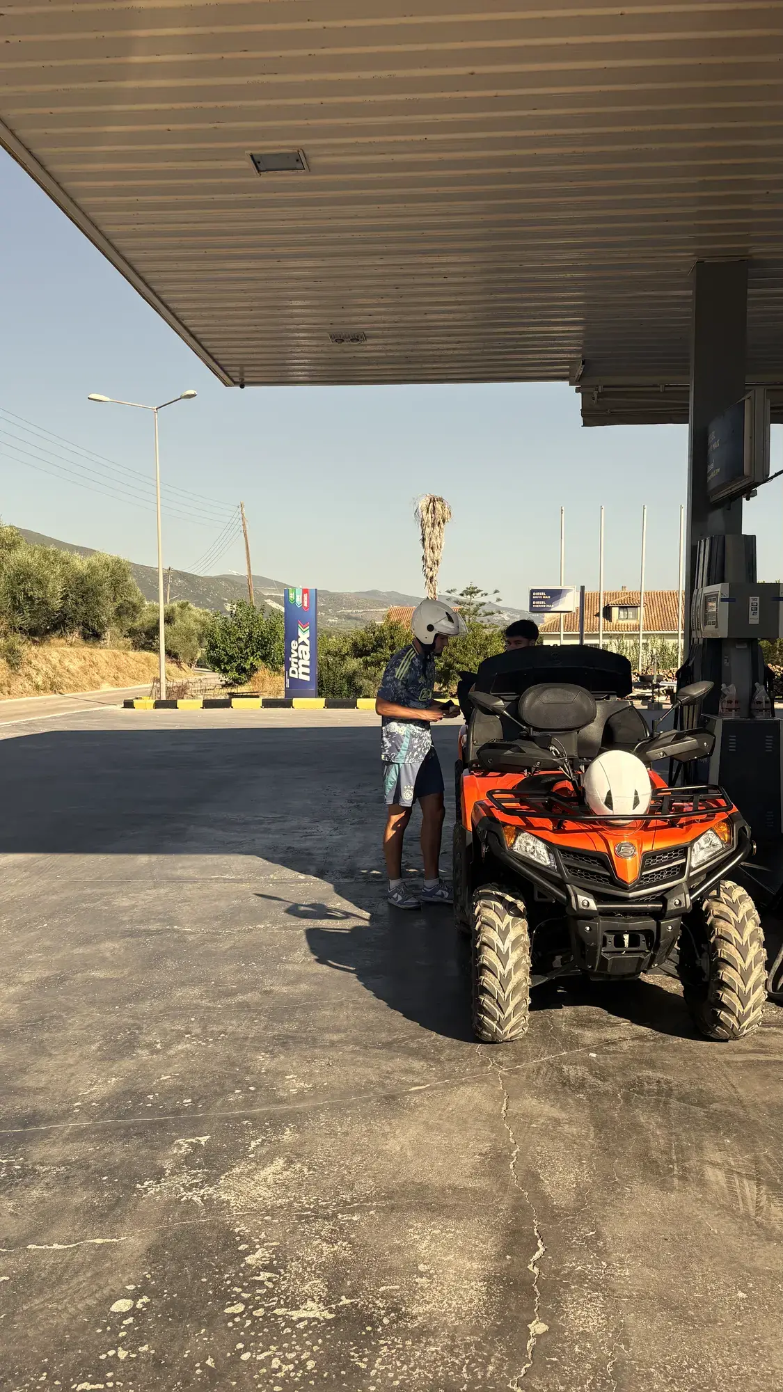 Jongen met helm naast een gehuurde oranje quad bij een tankstation op Zakynthos, met olijfbomen en heuvels op de achtergrond