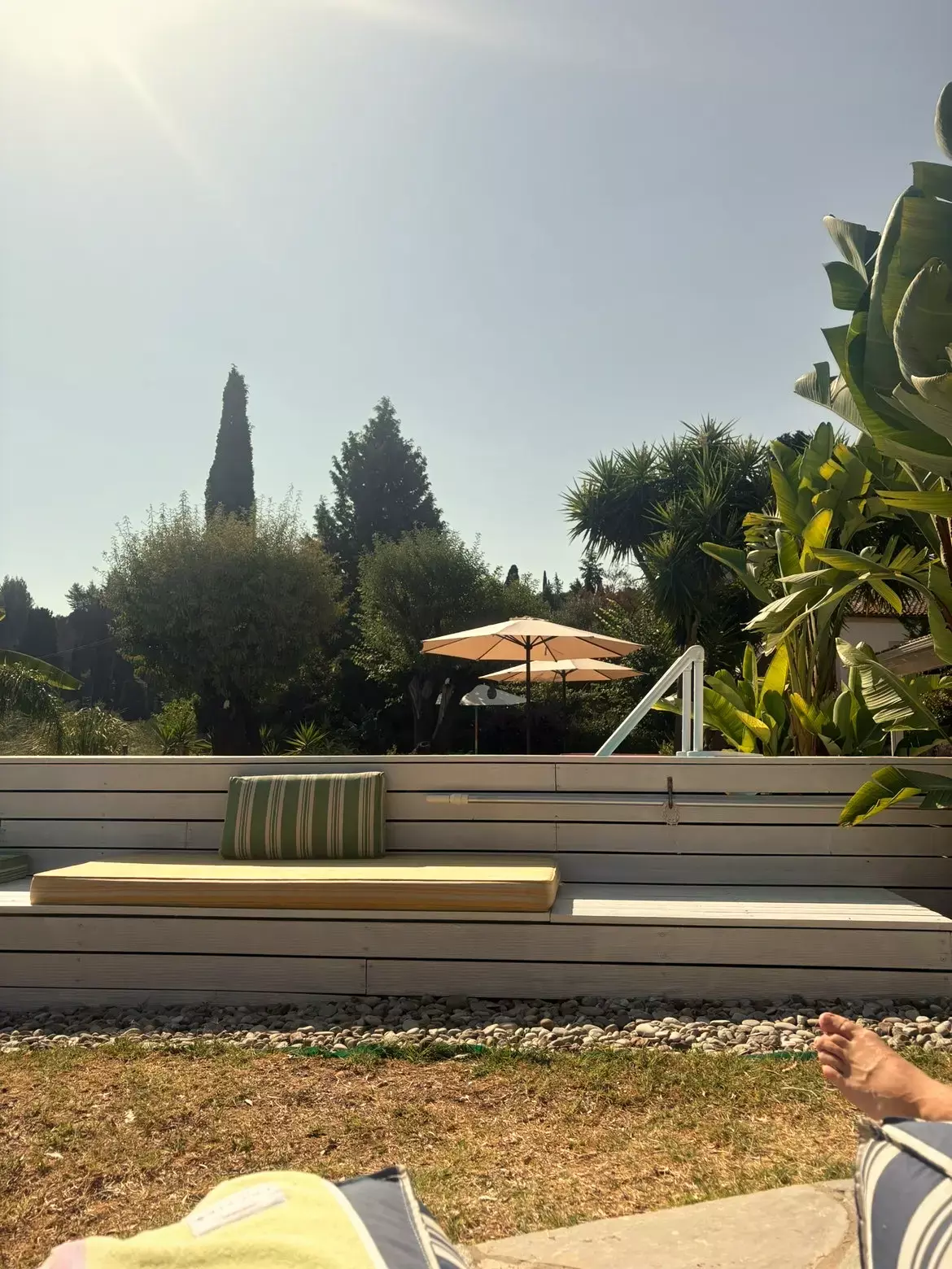 Houten loungebank met kussens in een zonnige villatuin op Corfu met parasols, palmen en cypressen