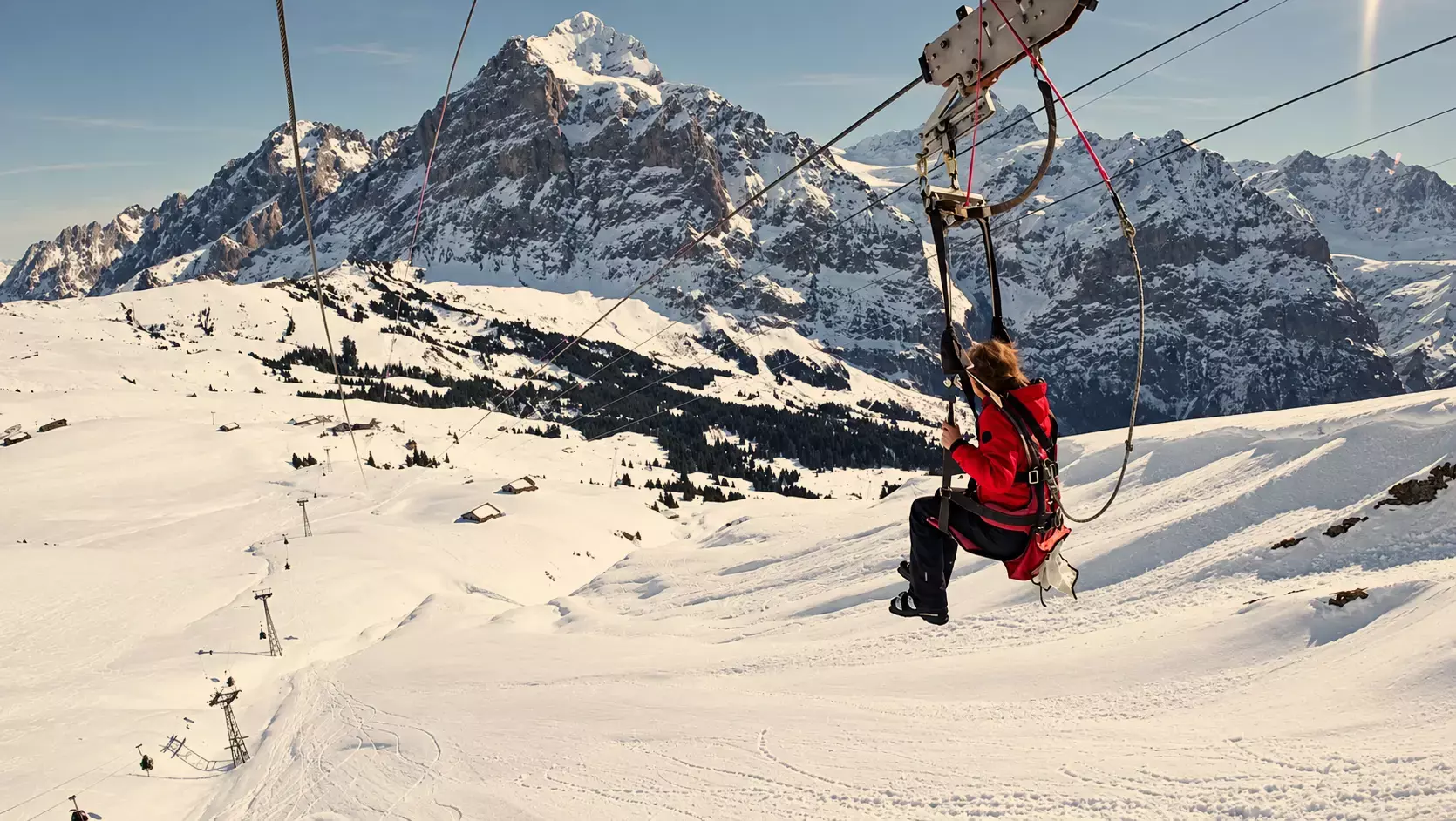 Iemand in een rode jas zoeft met de First Flyer zipline over de besneeuwde hellingen van Grindelwald First, met de Wetterhorn op de achtergrond