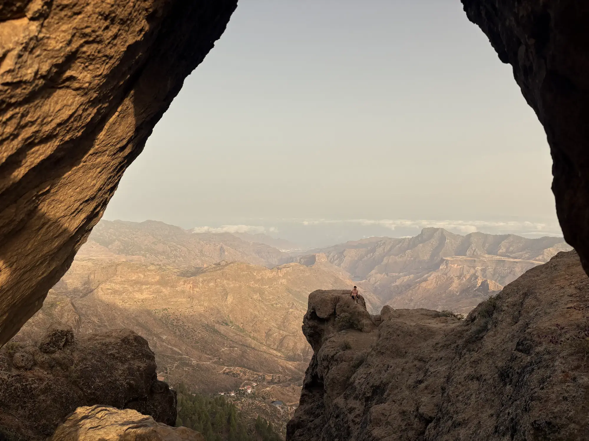 Uitzicht door een rotsboog op een ruig bergdal, met een persoon op een rotspunt in het gouden uur Roque Nublo
