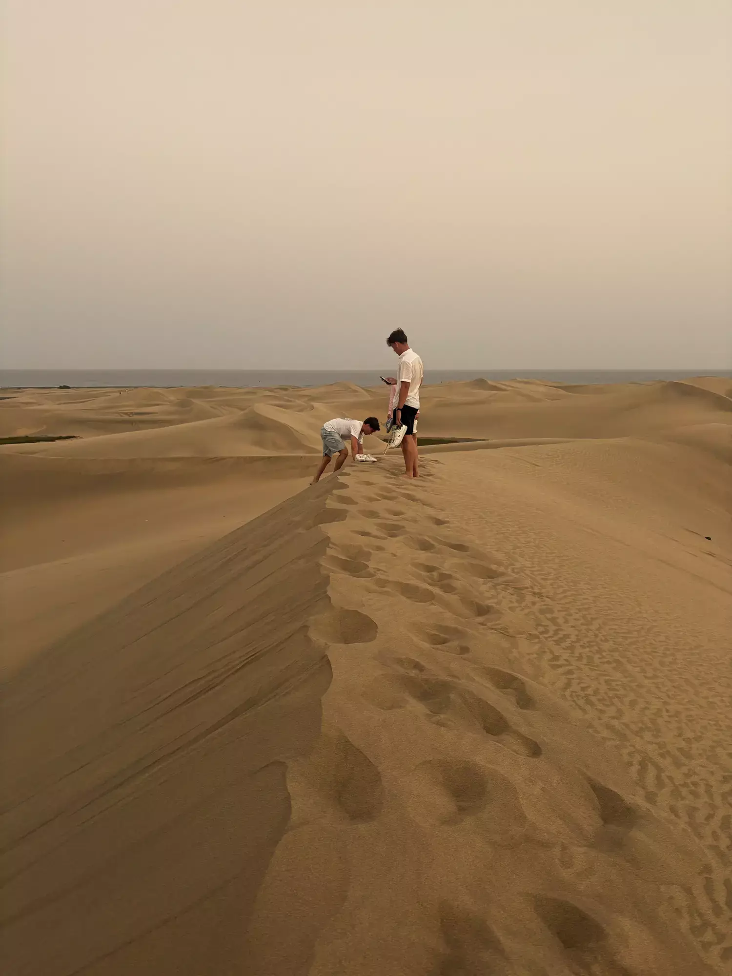 Twee mannen op een duinrug vol voetstappen in de duinen van Maspalomas, met de zee op de achtergrond