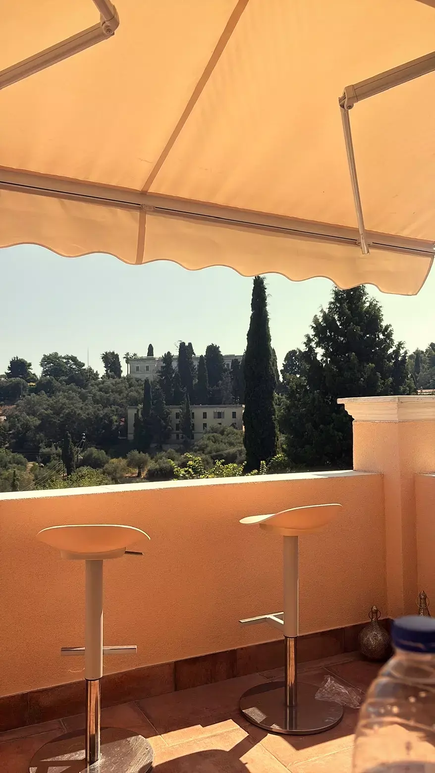 Balkon met witte barkrukken onder een oranje zonnescherm, met uitzicht op de cypressen van Corfu