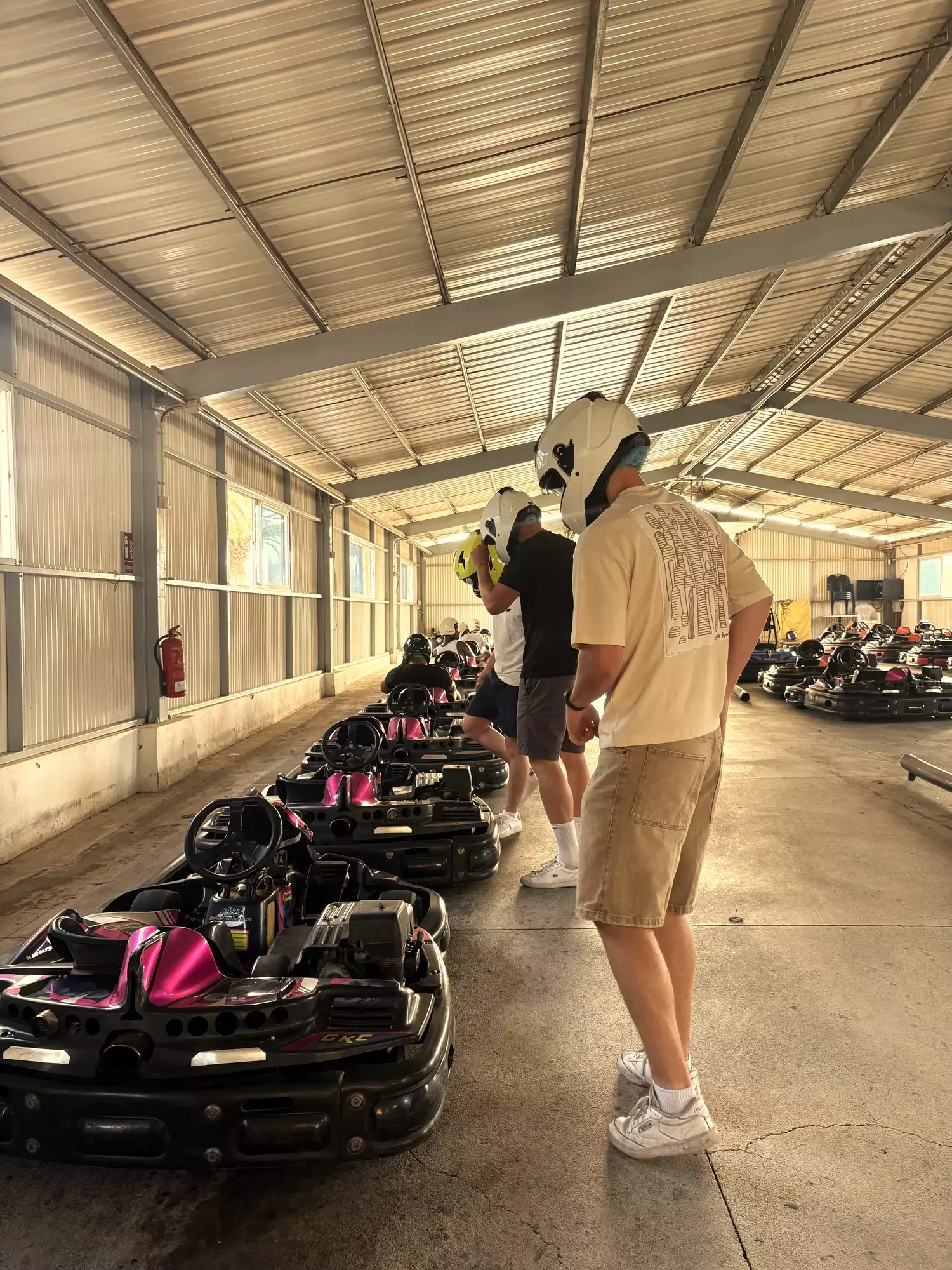 Karters met witte helmen staan klaar naast een rij roze-zwarte karts onder een overkapping