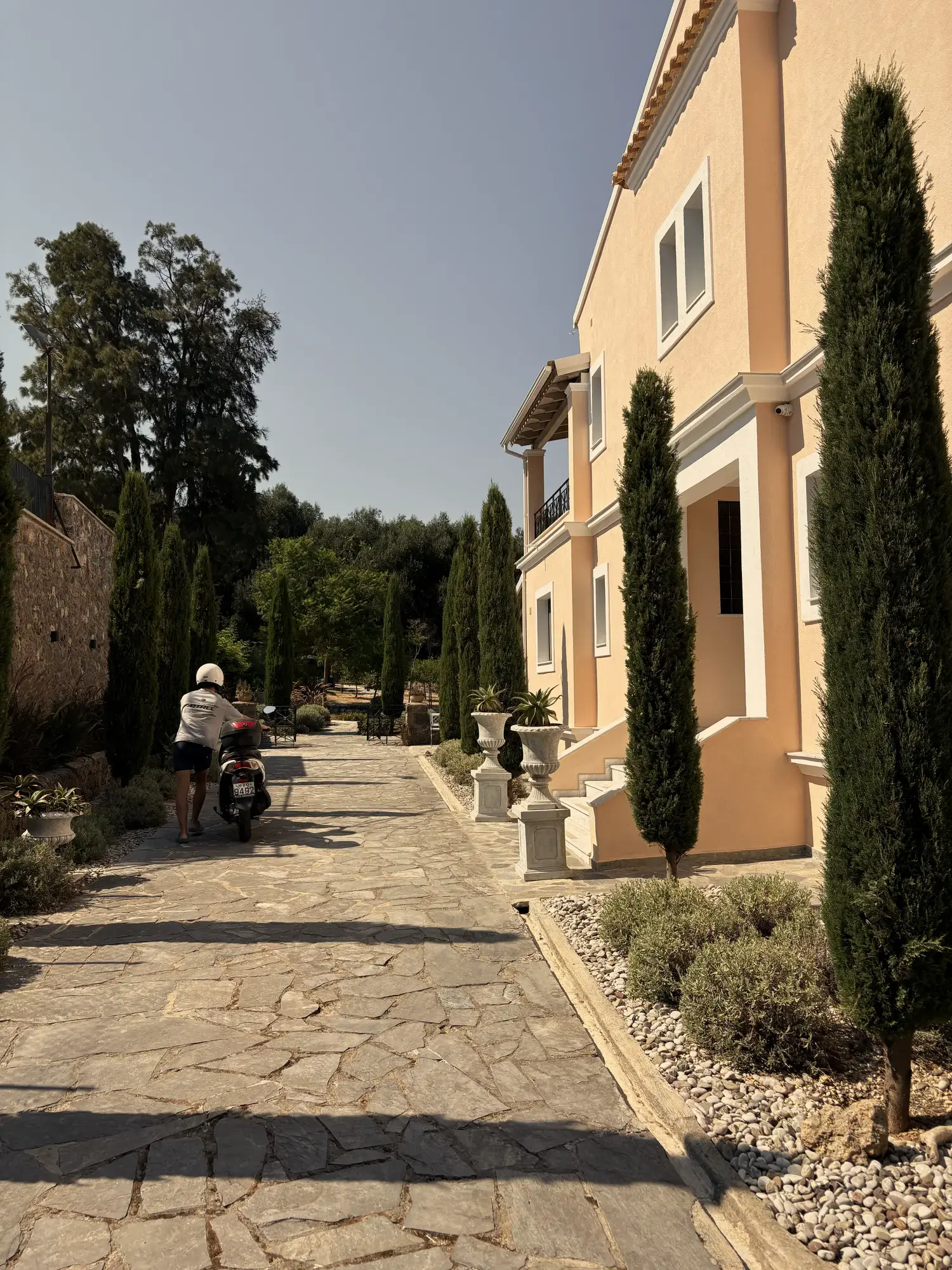 Zonnige natuurstenen oprit langs een zalmroze villa met cipressen, en iemand met een scooter