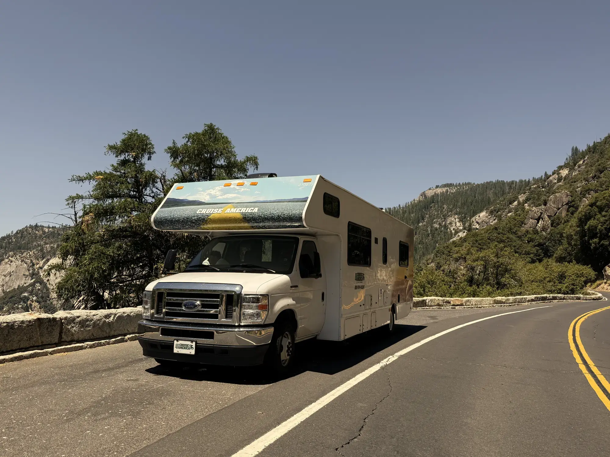 Witte Cruise America-camper op een bochtige bergweg met beboste rotshellingen onder een strakblauwe lucht