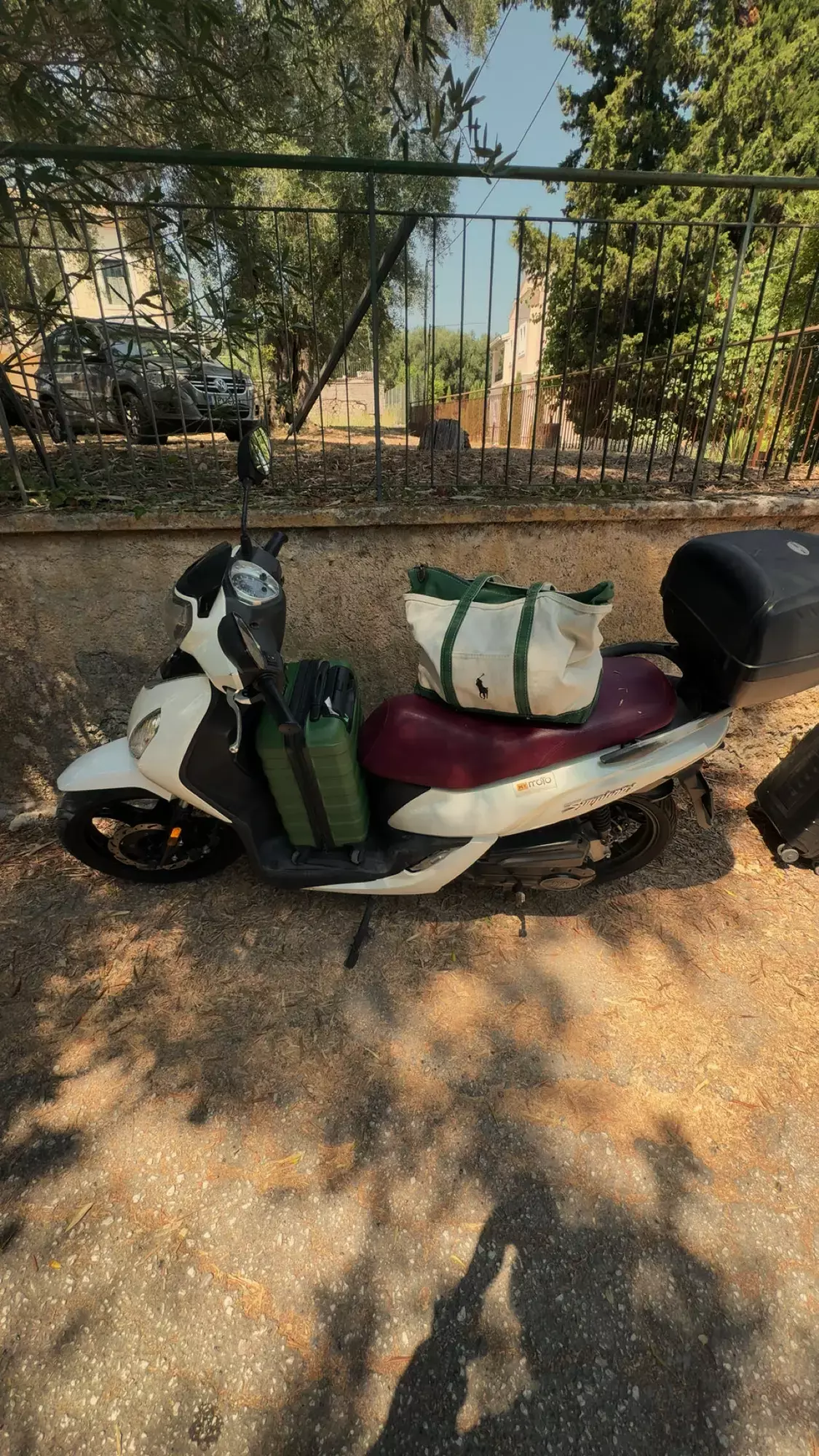 Witte scooter met groene tassen en topkoffer geparkeerd langs een hek op Corfu