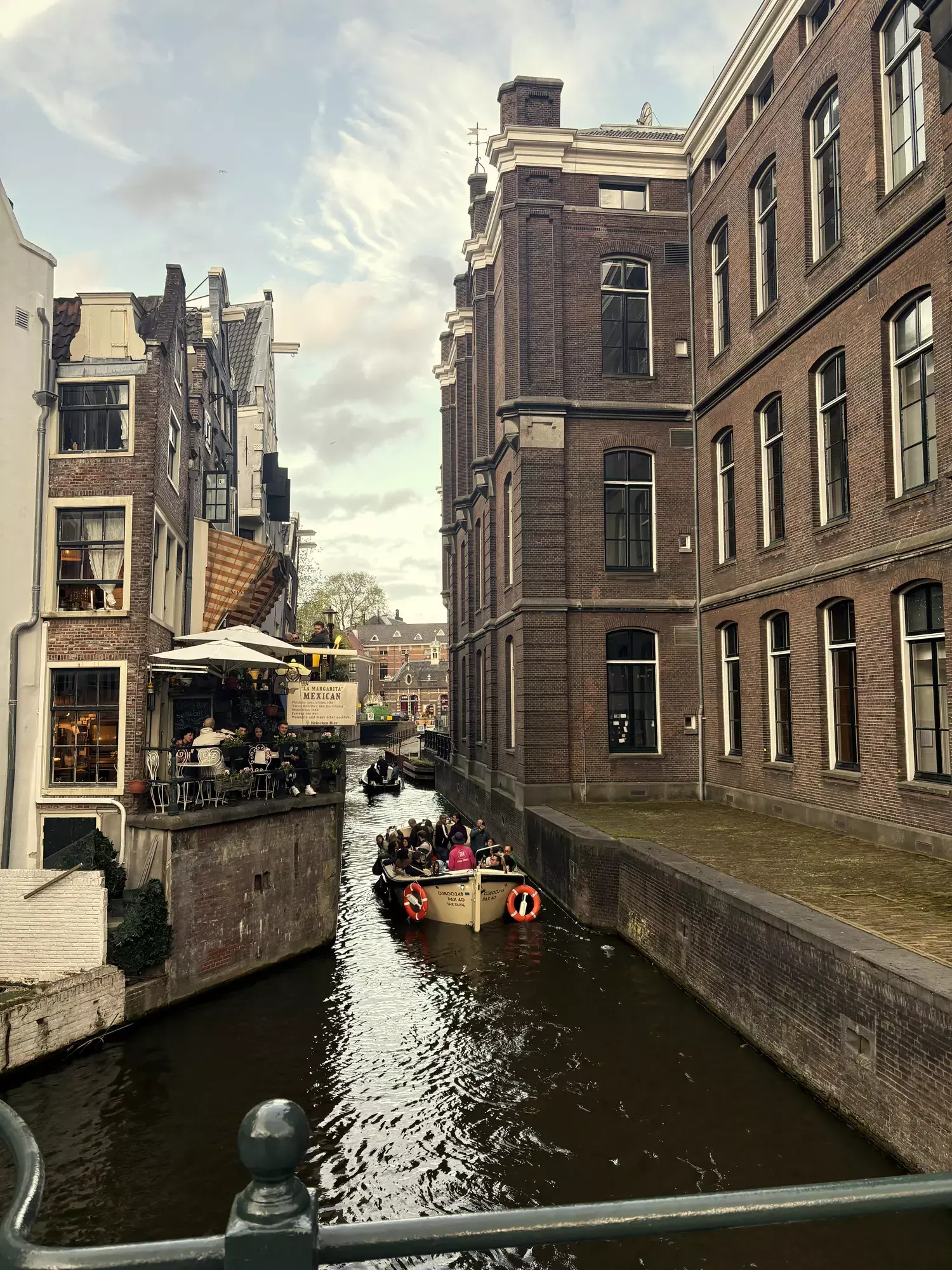 Rondvaartboot vaart door een smalle gracht in Amsterdam tussen bakstenen gebouwen en een terras aan het water