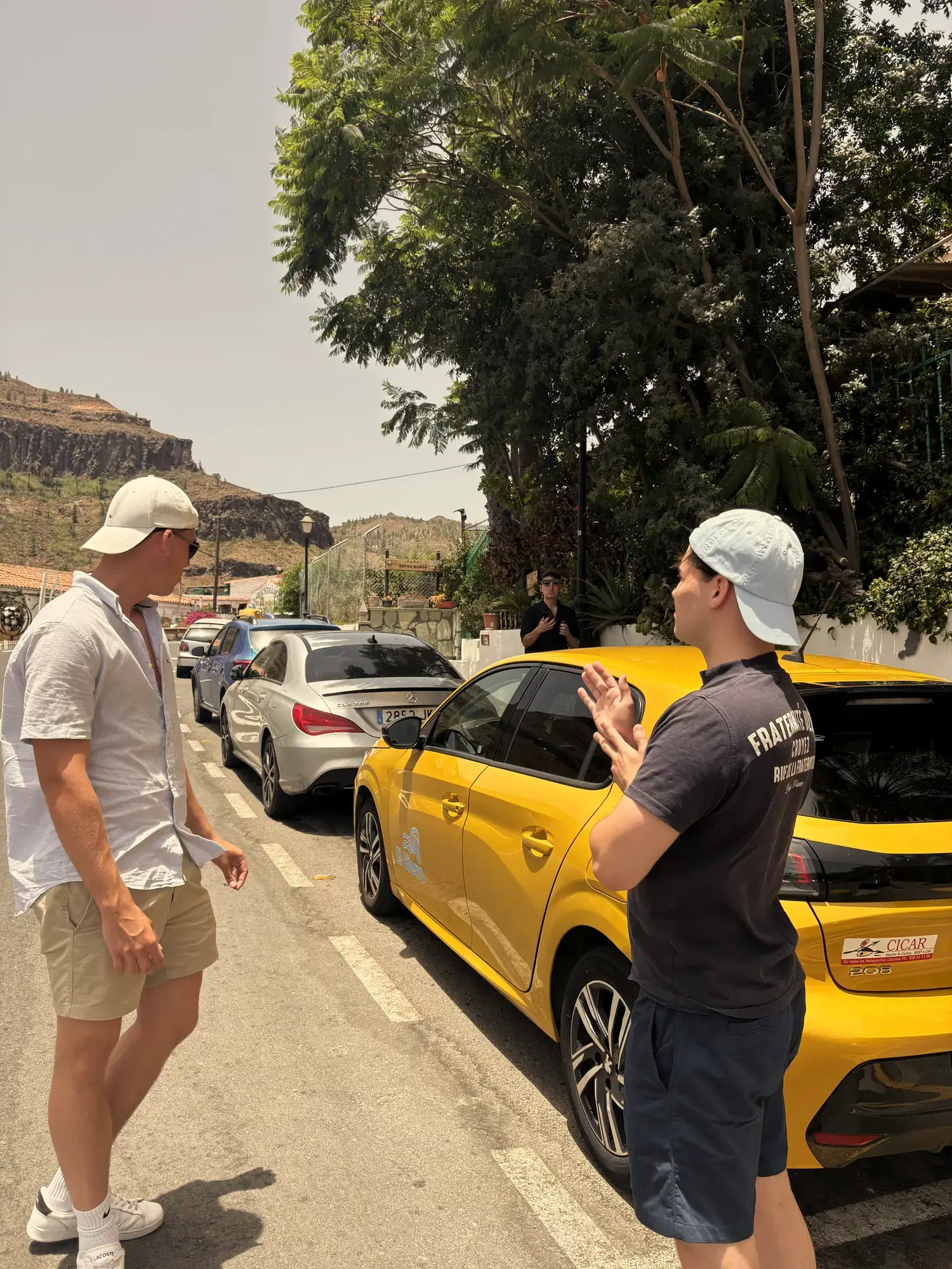 Twee mannen in gesprek naast een gele Peugeot 208 huurauto in een straat met rotsige heuvels op Gran Canaria