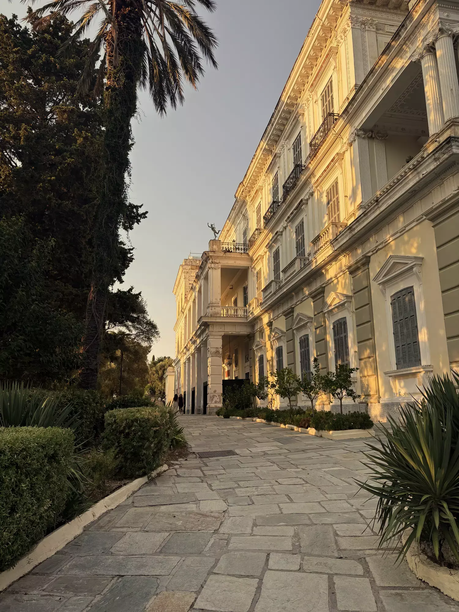Lichtgele gevel van het Achilleion-paleis langs een stenen wandelpad met palmen
