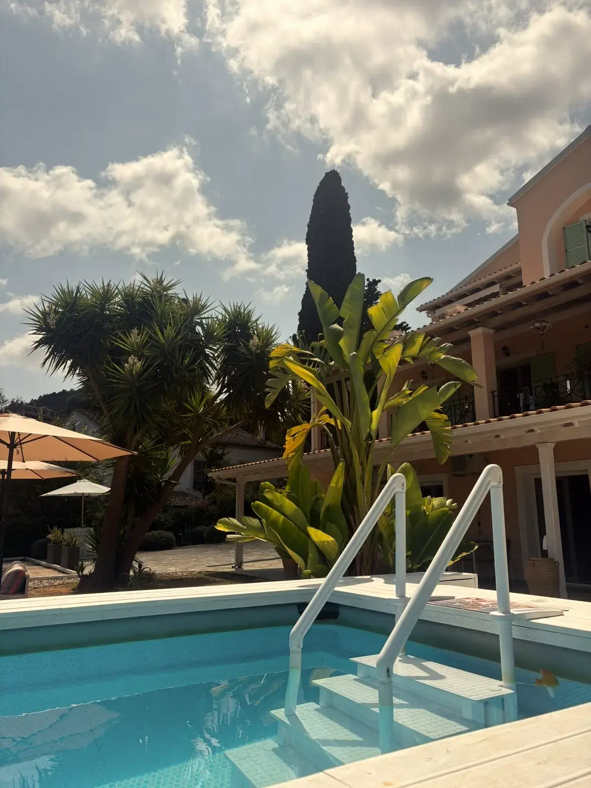 Zwembad met trapleuning bij een zalmroze villa op Corfu met palmen, bananenplanten en een cypres